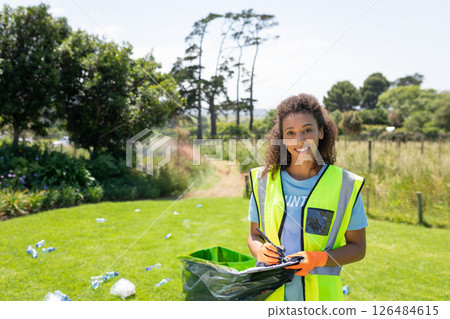 Smiling woman in safety vest cleaning park, holding trash bag and clipboard, copy space 126484615