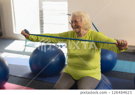 Senior woman exercising with resistance band on yoga ball, smiling in fitness class 126484623