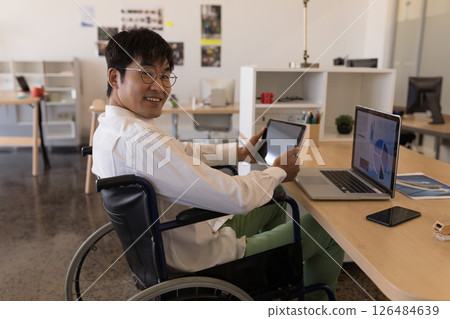 Man in wheelchair using tablet and laptop at office, smiling confidently 126484639