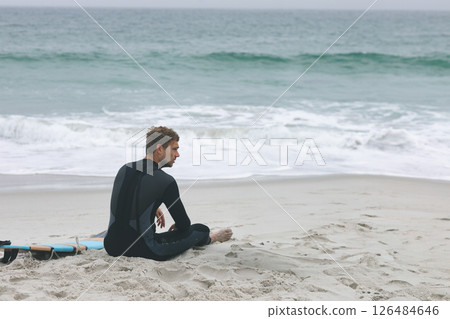 Surfer in wetsuit sitting on sandy beach, gazing at ocean waves, copy space 126484646