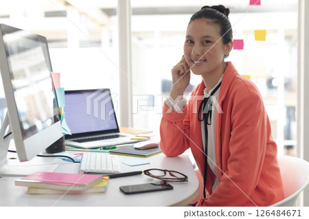 Smiling businesswoman in orange blazer working at desk with computer and smartphone, copy space 126484671