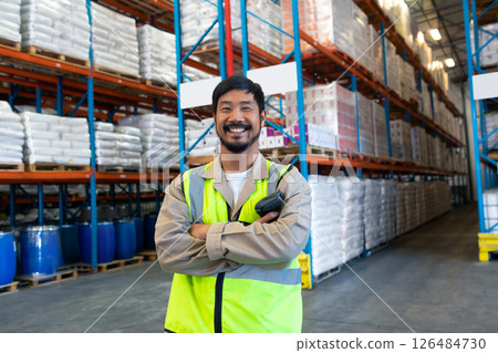 In warehouse, worker standing confidently with arms crossed among stacked shelves 126484730