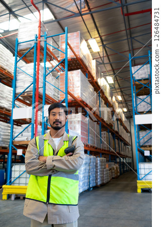 Warehouse worker in safety vest standing confidently in front of tall shelves, copy space 126484731