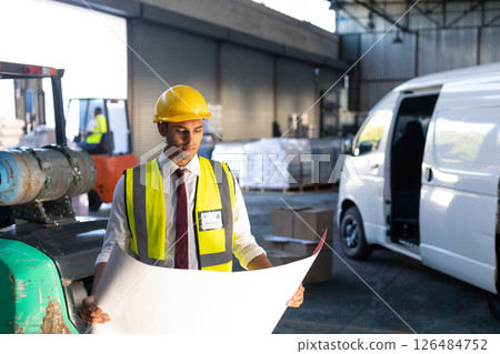 Engineer in safety vest reviewing blueprints in busy warehouse with delivery van, copy space 126484752