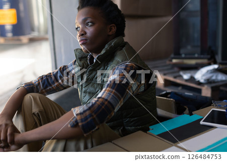 Contemplative woman in warehouse sitting among boxes, wearing plaid shirt Contemplative woman in warehouse sitting among boxes, wearing plaid shirt 126484753