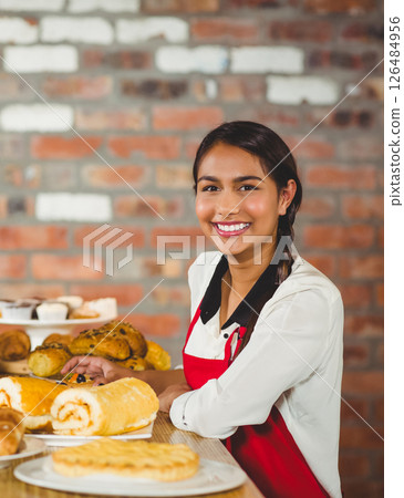 Smiling woman in red apron presenting fresh pastries at bakery counter Smiling woman in red apron presenting fresh pastries at bakery counter 126484956