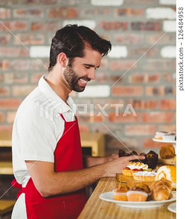 Barista in red apron arranging pastries with smile at cozy café counter Barista in red apron arranging pastries with smile at cozy café counter 126484958