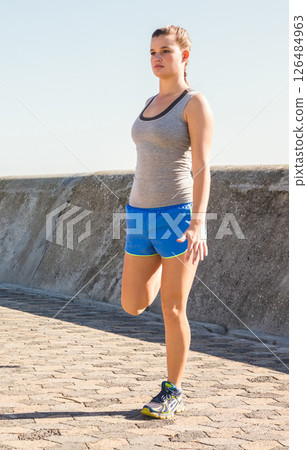 Woman stretching leg on sunny promenade, preparing for morning exercise routine 126484963