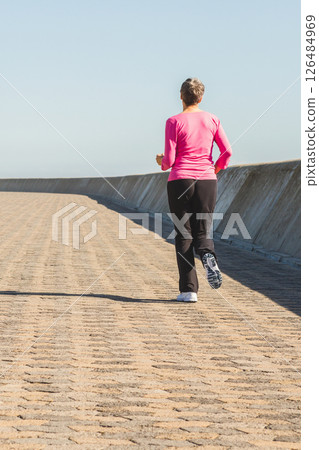 Woman jogging on sunny promenade wearing pink top and black pants, copy space 126484969