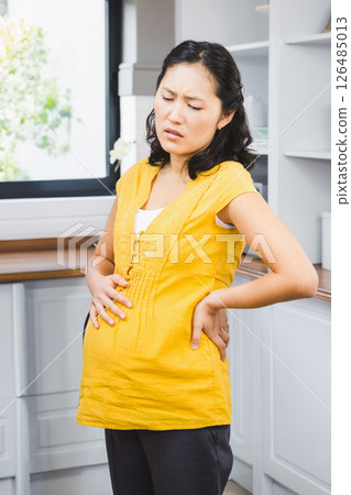 Pregnant Asian woman in kitchen experiencing discomfort, holding back and belly 126485013