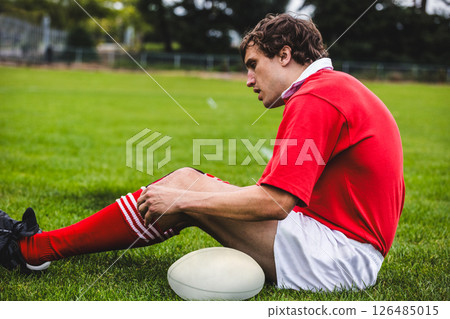 Rugby player in red jersey resting on field with rugby ball 126485015