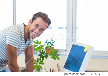 Smiling man leaning on desk in office, working with computer and plant Smiling man leaning on desk in office, working with computer and plant 126485113