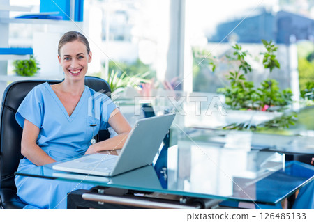 Smiling nurse in scrubs working on laptop at modern medical office 126485133