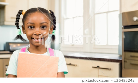 Smiling African American girl holding book in kitchen, ready for school, copy space 126485242