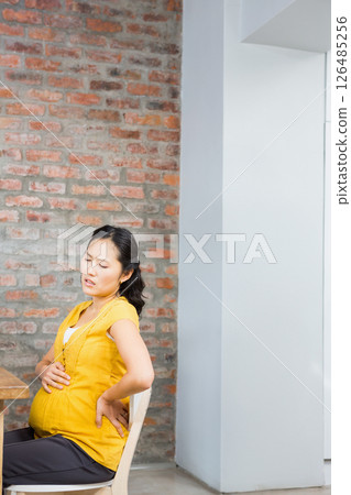 Pregnant Asian woman sitting at home, experiencing back pain, holding belly, copy space 126485256