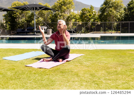 Senior man meditating on yoga mat by pool, enjoying peaceful outdoor setting, copy space 126485294