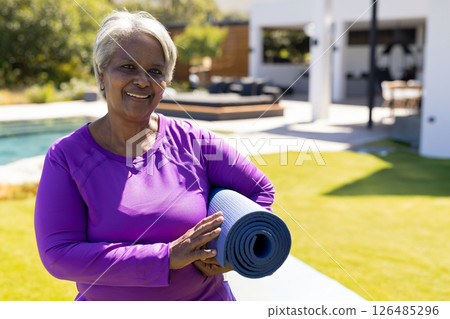 Senior woman holding yoga mat, smiling by poolside, enjoying outdoor relaxation, copy space 126485296