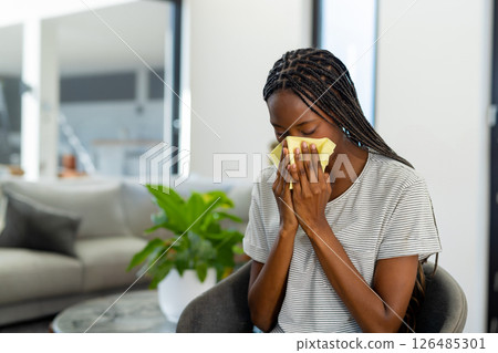 Woman sneezing into tissue at home, sitting on chair, feeling unwell, copy space 126485301