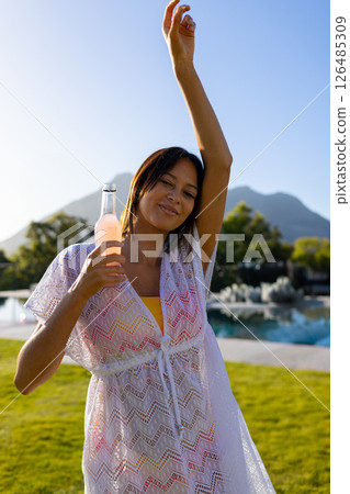 Smiling woman enjoying refreshing drink by pool, raising arm in relaxation, copy space Smiling woman enjoying refreshing drink by pool, raising arm in relaxation, copy space 126485309