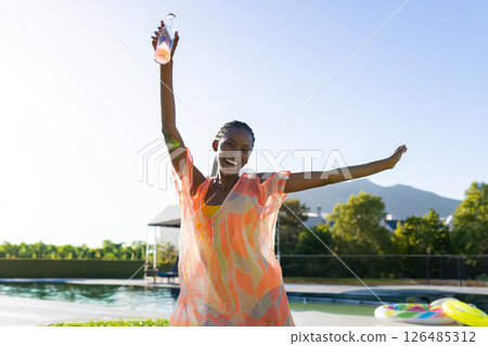 Joyful woman in colorful dress celebrating by pool, holding refreshing drink, copy space 126485312