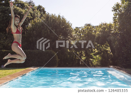 Woman in red bikini joyfully jumping into pool at outdoor party, copy space 126485335
