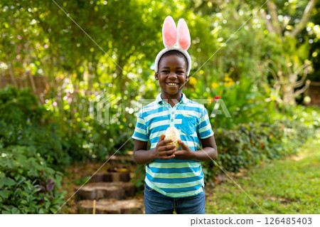 Smiling boy wearing bunny ears holding toy outdoors in lush garden, copy space Smiling boy wearing bunny ears holding toy outdoors in lush garden, copy space 126485403