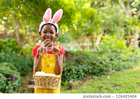Smiling child wearing bunny ears holding basket with eggs in garden, copy space 126485404