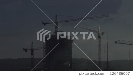 Construction cranes towering over high-rise building under dark, cloudy sky 126486007