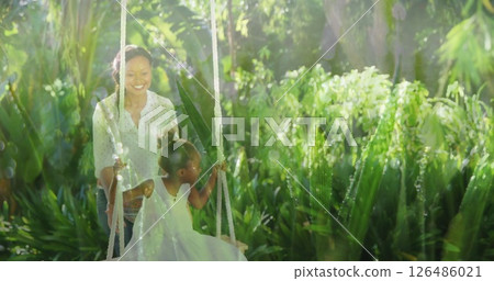 Pushing daughter on swing in lush garden, African American mother smiling joyfully Pushing daughter on swing in lush garden, African American mother smiling joyfully 126486021