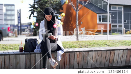 Young woman in hijab using tablet outdoors, engaging with social media icons 126486114