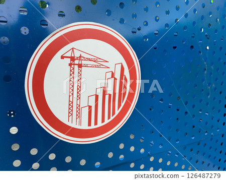 Red construction themed warning sign with cranes and skyscrapers printed on perforated blue metal fencing, symbolizing active development zone and modern urban growth initiatives 126487279