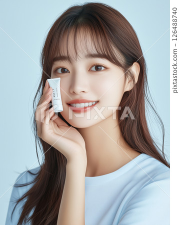 Korean woman smiling while holding skincare product in a light blue background during a beauty photoshoot Korean woman smiling while holding skincare product in a light blue background during a beauty photoshoot 126488740