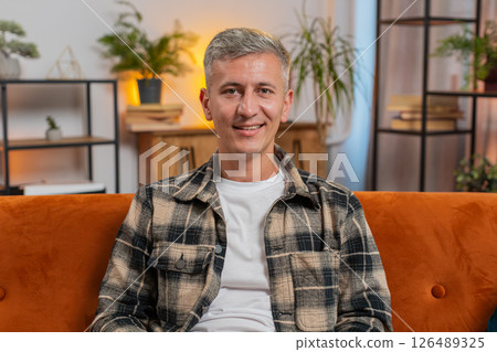 Portrait of happy young Caucasian man sitting on orange sofa looking at camera and smiling at home 126489325