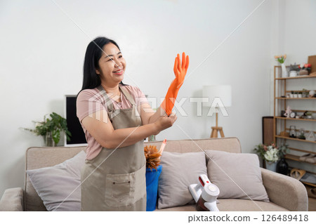 Ready for Cleaning. A woman smiles while putting on orange gloves in her living room. Ready for Cleaning. A woman smiles while putting on orange gloves in her living room. 126489418