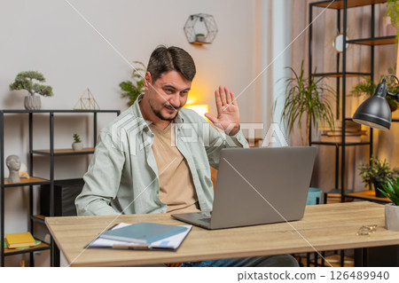 Smiling young man freelancer having video call via laptop sitting at home office desk in living room Smiling young man freelancer having video call via laptop sitting at home office desk in living room 126489940