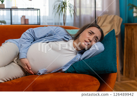 Upset ill young pregnant woman feeling sick suffering from abdominal belly pain lying on home sofa Upset ill young pregnant woman feeling sick suffering from abdominal belly pain lying on home sofa 126489942
