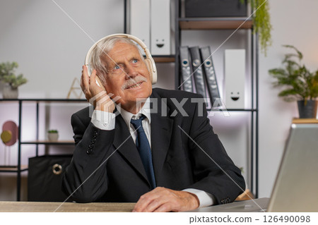 Overjoyed senior businessman sitting at home office desk listening to music through headphones Overjoyed senior businessman sitting at home office desk listening to music through headphones 126490098