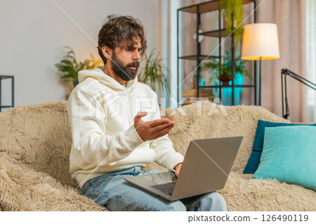 Upset young Indian man freelancer using laptop engages in heated conversations sitting on sofa 126490119