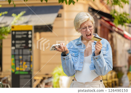 Upset young woman with short blond hair using smartphone and credit card on urban city street 126490150