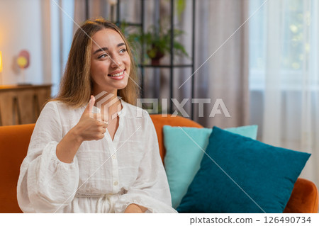Portrait of excited smiling young woman showing double thumbs up sitting on sofa couch at home Portrait of excited smiling young woman showing double thumbs up sitting on sofa couch at home 126490734