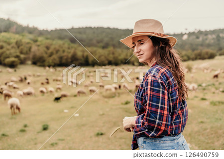 A woman wearing a straw hat stands in a field of sheep 126490759