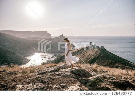 woman stands on a rocky hill overlooking a body of water. She is wearing a white dress and she is in a state of joy or celebration. Concept of freedom and happiness. 126490777