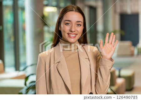 Happy young Caucasian businesswoman looking at camera and waves hand hello in office hotel lobby Happy young Caucasian businesswoman looking at camera and waves hand hello in office hotel lobby 126490785