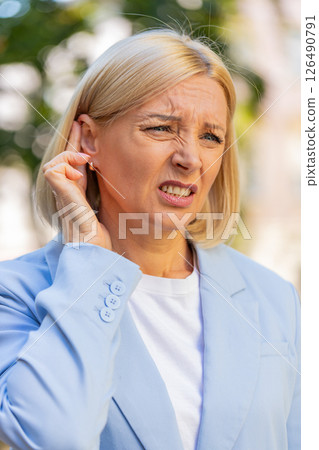 Sick mature businesswoman in formal suit suffers from ear pain while standing on city town street 126490791