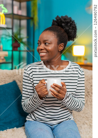 African American young woman relaxing at home on couch, holding hot tea cup, dreaming, smiling 126490792