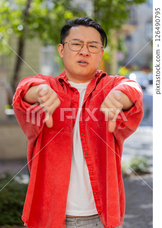 Upset Asian man showing thumbs down sign, disapproval, dissatisfied bad work, mistake in city Upset Asian man showing thumbs down sign, disapproval, dissatisfied bad work, mistake in city 126490795