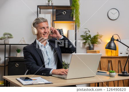 Happy young businessman in suit and wireless headphones enjoy music using laptop at home office desk Happy young businessman in suit and wireless headphones enjoy music using laptop at home office desk 126490820