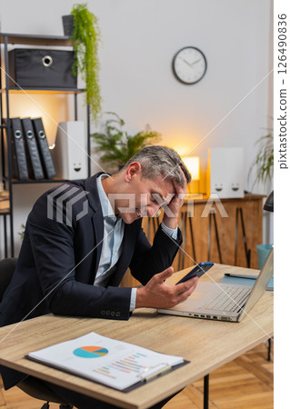Disappointed Caucasian young businessman reading bad news on cellphone sitting at home office table 126490836
