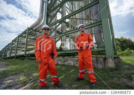 Two workers supervisor dressed in orange safety suits and white helmets observing pipeline project at construction site. engineer at industrial tube oil petrochemical. 126490873
