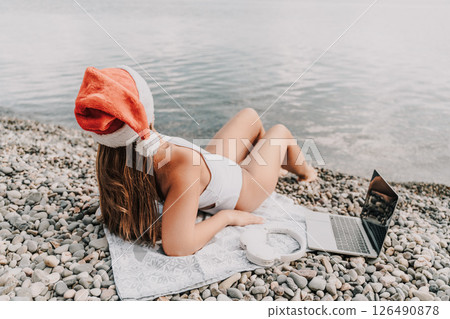 A woman is laying on the beach with a laptop and headphones on her head. She is wearing a red Santa hat. A woman is laying on the beach with a laptop and headphones on her head. She is wearing a red Santa hat. 126490878